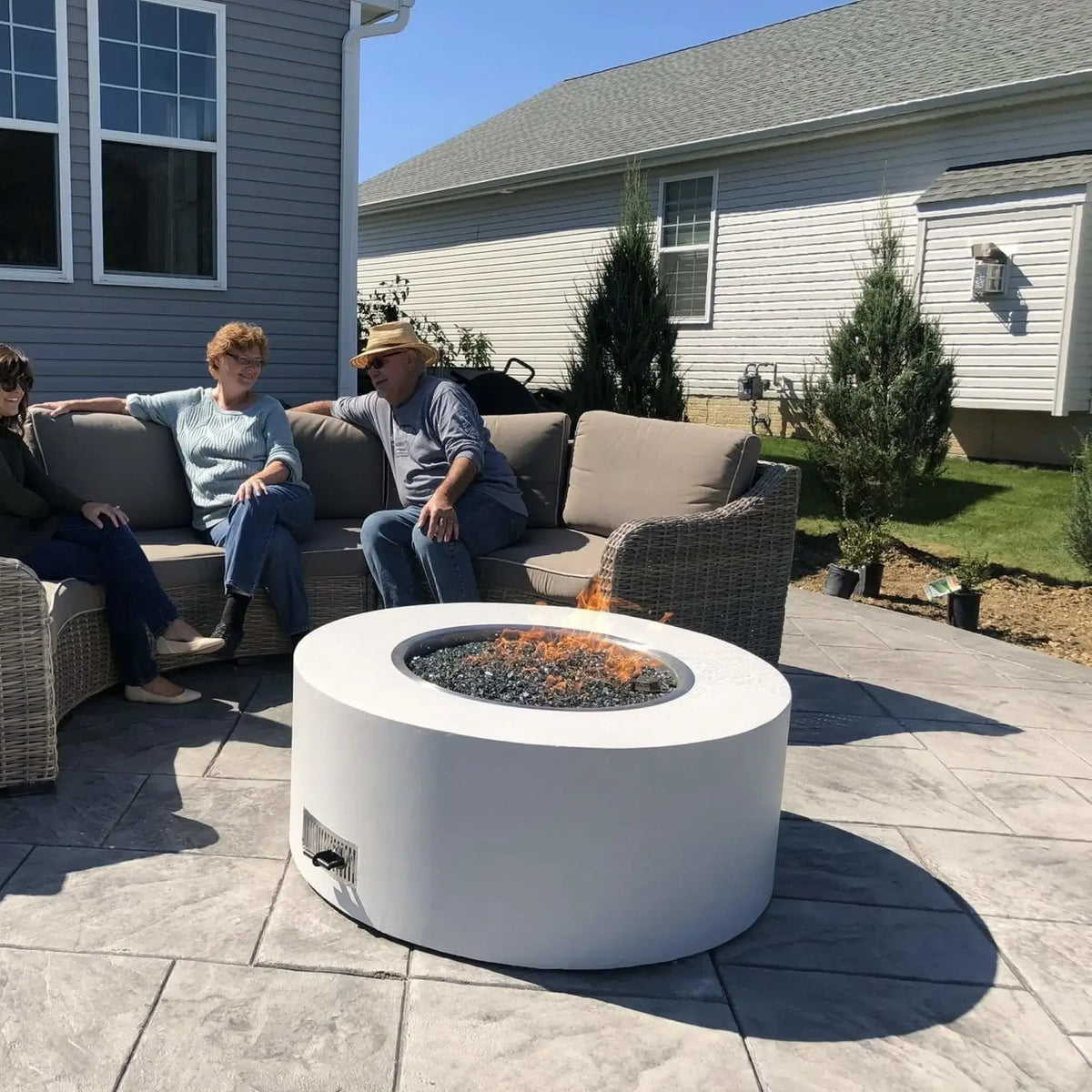 Round white concrete gas fire pit with black fire glass burning bright flames, centered on a stamped concrete patio where three adults relax on a wicker sectional, creating a cozy backyard gathering space in a suburban setting.