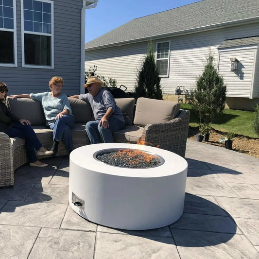 Round white concrete gas fire pit with black fire glass burning bright flames, centered on a stamped concrete patio where three adults relax on a wicker sectional, creating a cozy backyard gathering space in a suburban setting.