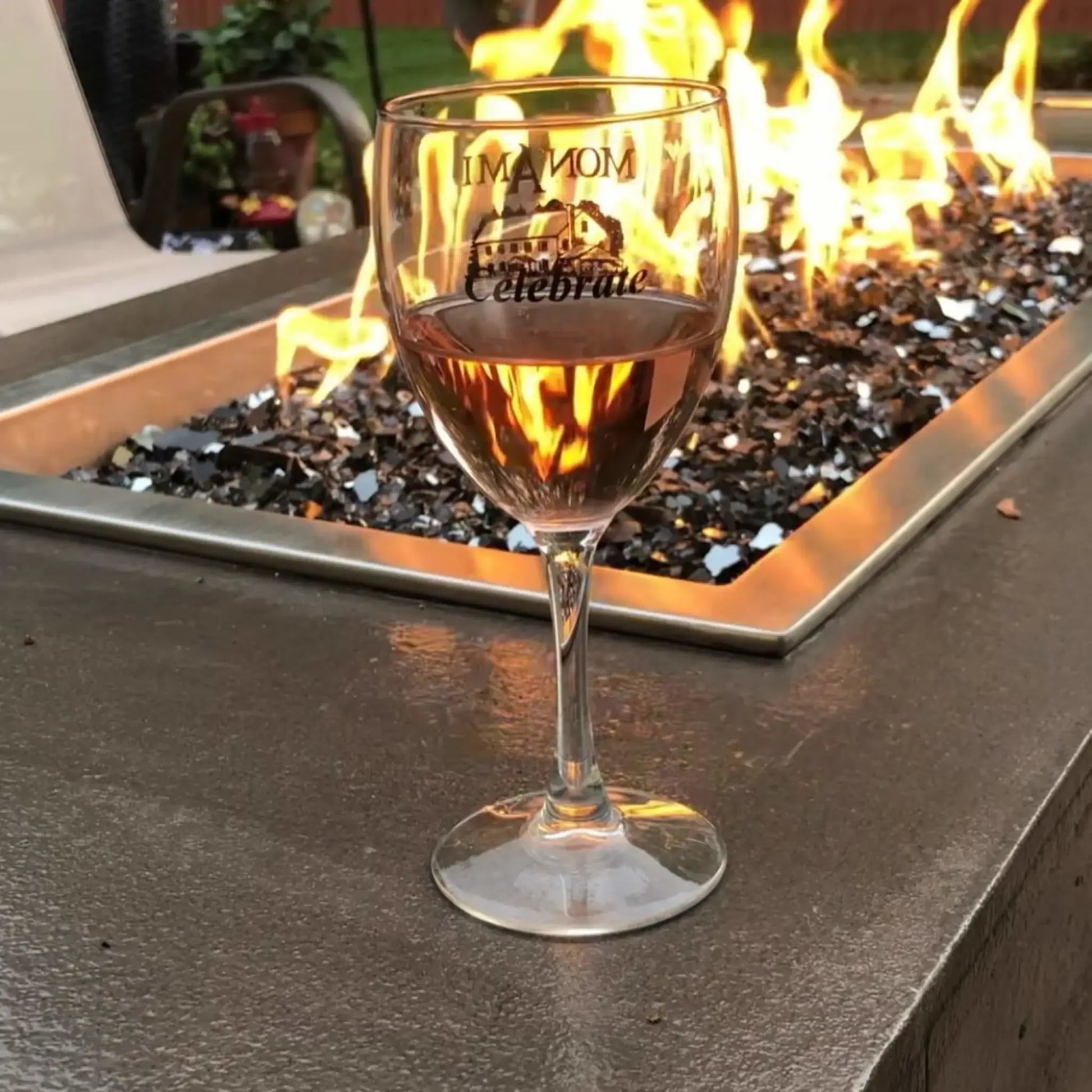 Close-up of a wine glass filled with rosé placed beside a rectangular fire pit burner filled with glossy black fire glass, reflecting flickering flames and creating a warm, intimate outdoor ambiance.