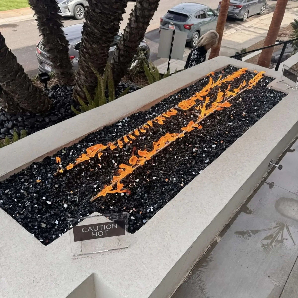 Long rectangular concrete fire feature filled with black fire glass and dual linear flame rows, installed in a landscaped front yard with palm trees and street view, including a small “caution hot” sign near the burner.