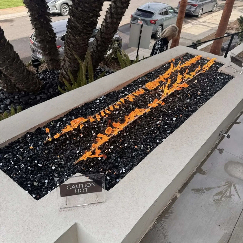 Long rectangular concrete fire feature filled with black fire glass and dual linear flame rows, installed in a landscaped front yard with palm trees and street view, including a small “caution hot” sign near the burner.