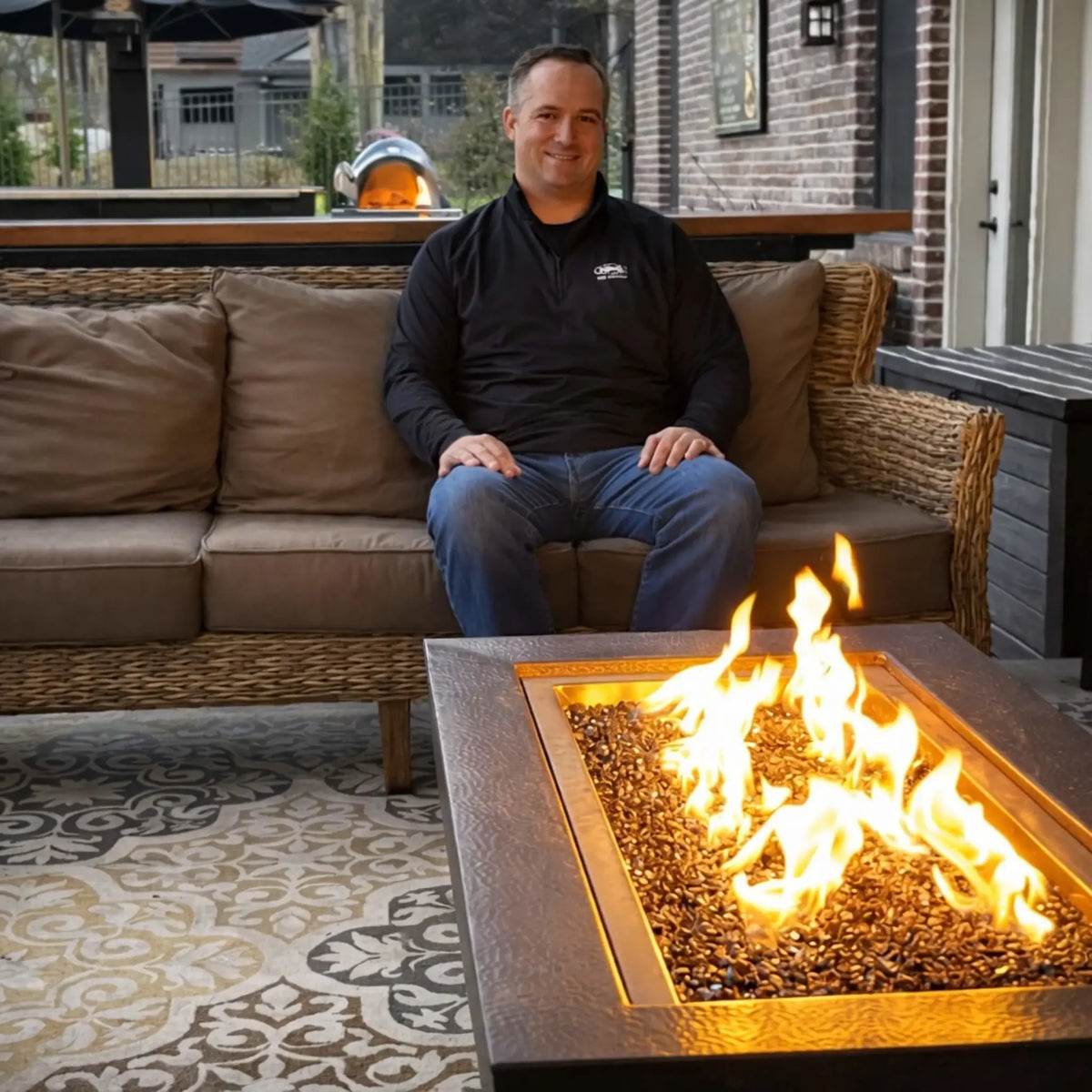 Man seated on a wicker outdoor sofa beside a square gas fire pit table filled with black fire glass, with bright flames illuminating a covered patio featuring patterned flooring and a built-in outdoor kitchen in the background.