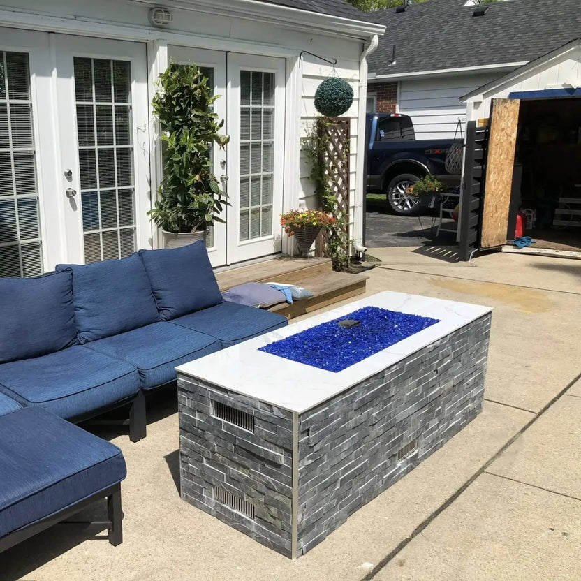 Modern rectangular outdoor fire pit table with gray stacked stone base and white top, filled with vibrant cobalt blue fire glass, positioned beside a blue cushioned sectional on a backyard patio near a home with French doors.