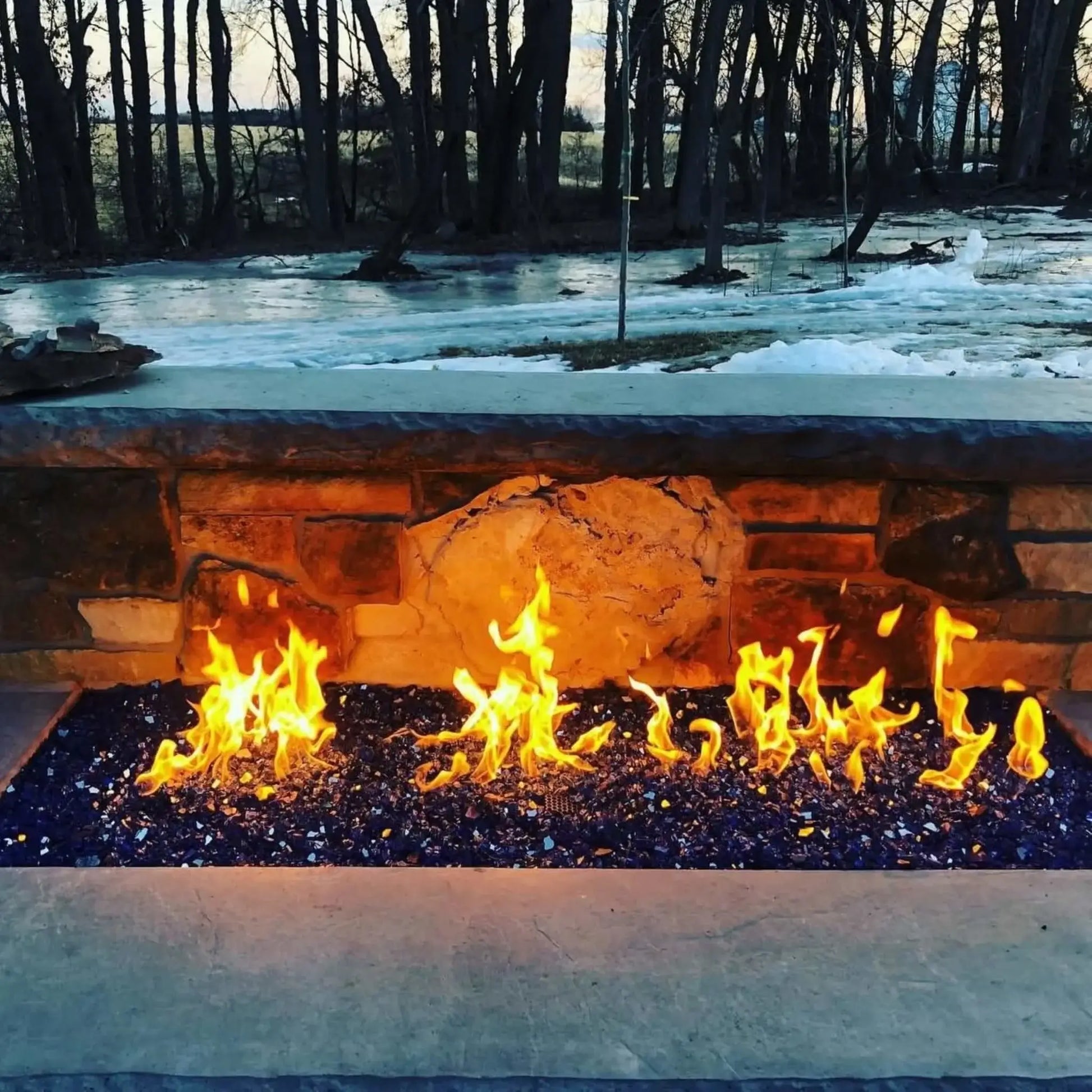 Linear outdoor fire feature filled with cobalt blue fire glass set into a stone wall, showcasing glowing flames against a winter landscape with snow-covered ground and trees, creating a striking contrast of warmth and cold.