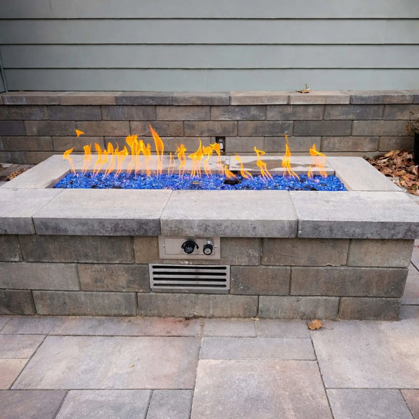 Rectangular outdoor gas fire pit built with gray stone blocks and a smooth concrete cap, featuring a long linear burner filled with vibrant cobalt blue fire glass producing bright flames, set against a neutral siding wall on a clean paved patio.