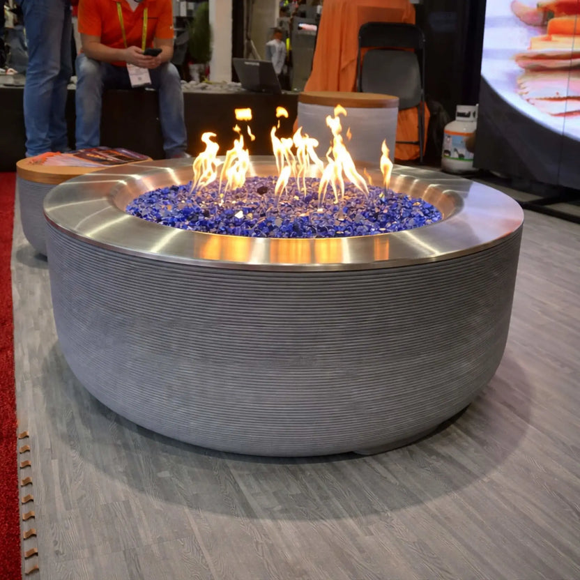Large round modern fire pit table with a brushed metal rim and textured base, filled with cobalt blue fire glass and lively flames, displayed indoors at a trade show booth with attendees and product displays in the background.