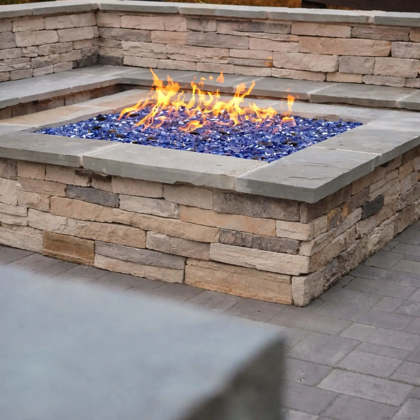 Square stone fire pit with a wide ledge and built-in seating wall, filled with cobalt blue fire glass and glowing flames, creating a cozy outdoor living space with natural stone textures and neutral patio pavers.