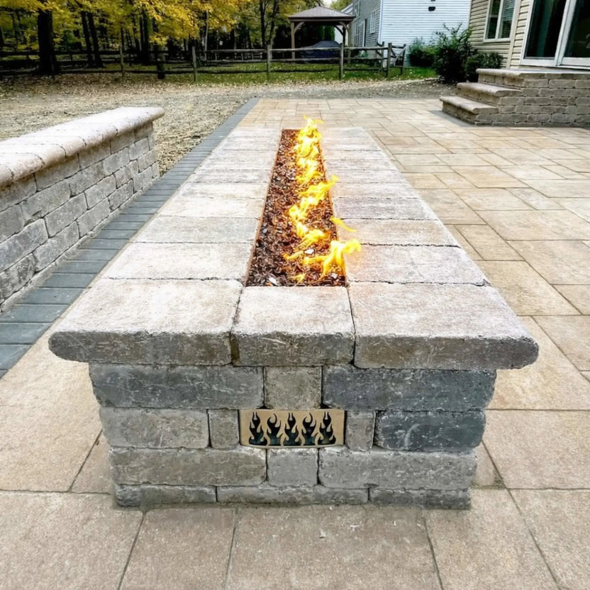 Long rectangular fire pit table constructed from gray stone pavers, featuring a narrow linear burner filled with dark reflective fire glass and bright flames, positioned on a spacious backyard patio surrounded by autumn trees and landscaping.