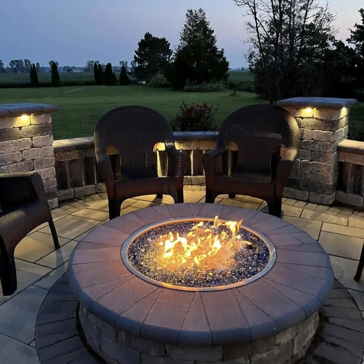 Round patio fire pit table with built-in circular burner filled with sparkling Pacific blue fire glass, surrounded by dark wicker chairs and stone columns with ambient lighting, creating a relaxing outdoor evening setting.