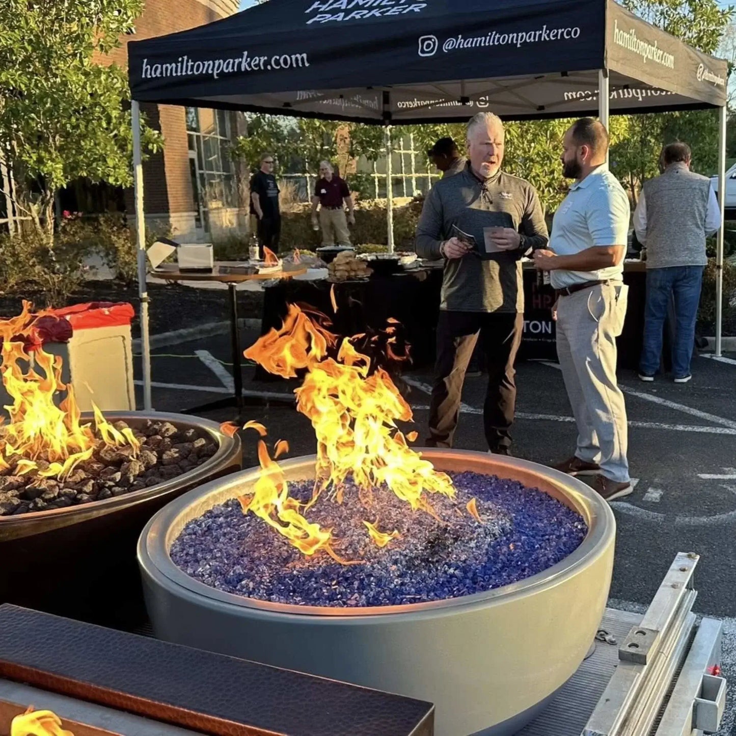 Outdoor event display featuring round fire bowls filled with Pacific blue fire glass and active flames, with people conversing under a branded canopy tent, highlighting a product demonstration in a casual gathering environment.