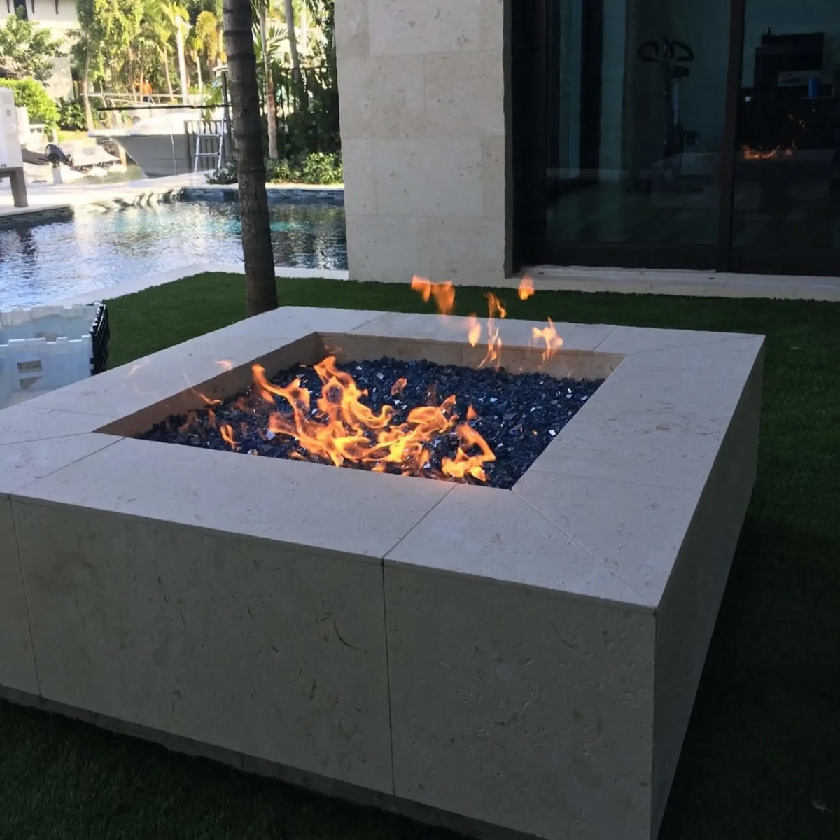 Square modern outdoor fire pit constructed from light beige stone with a recessed burner filled with dark reflective fire glass, producing warm flames beside a luxury poolside patio with tropical landscaping, water features, and a contemporary home exterior.