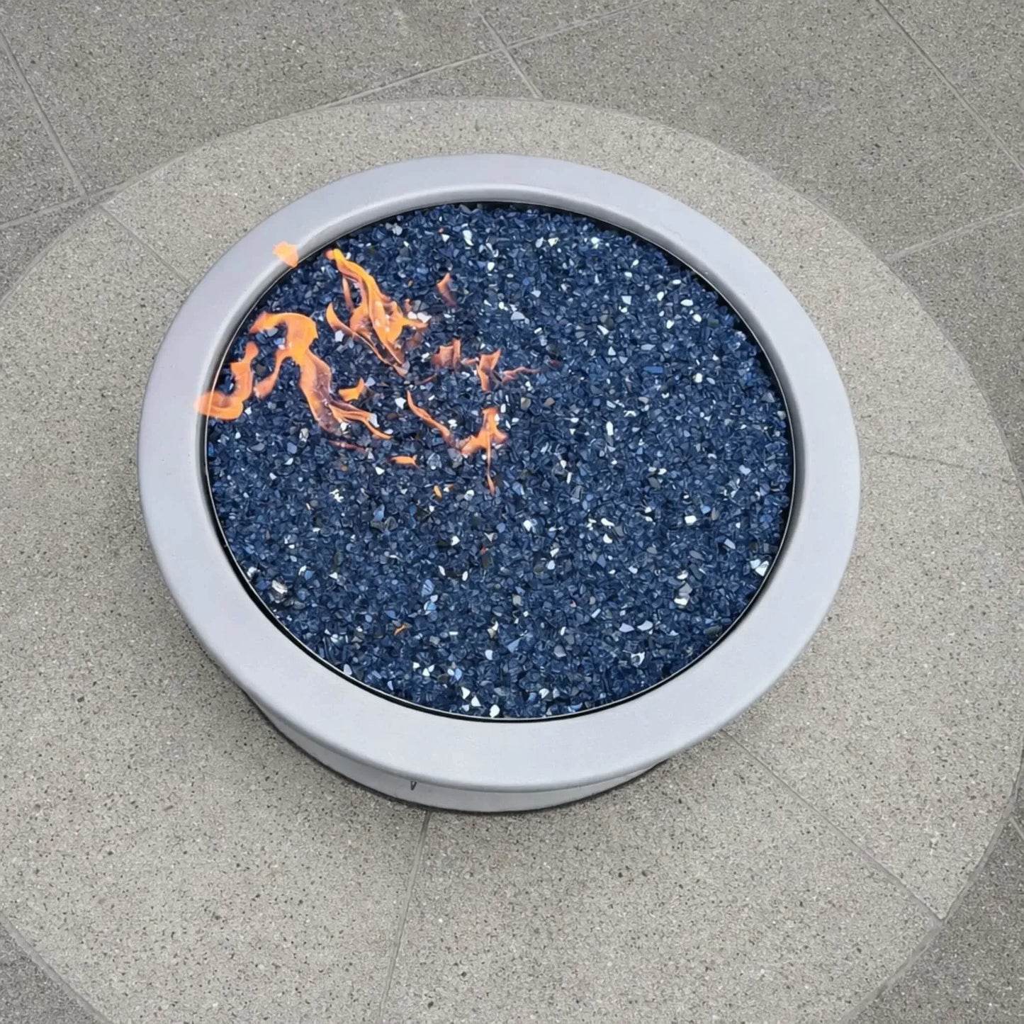 Top-down view of a circular fire pit burner filled with Pacific blue fire glass, set into a light concrete patio surface, with small orange flames flickering across the glass media, highlighting the clean, minimalist design and even burner distribution.