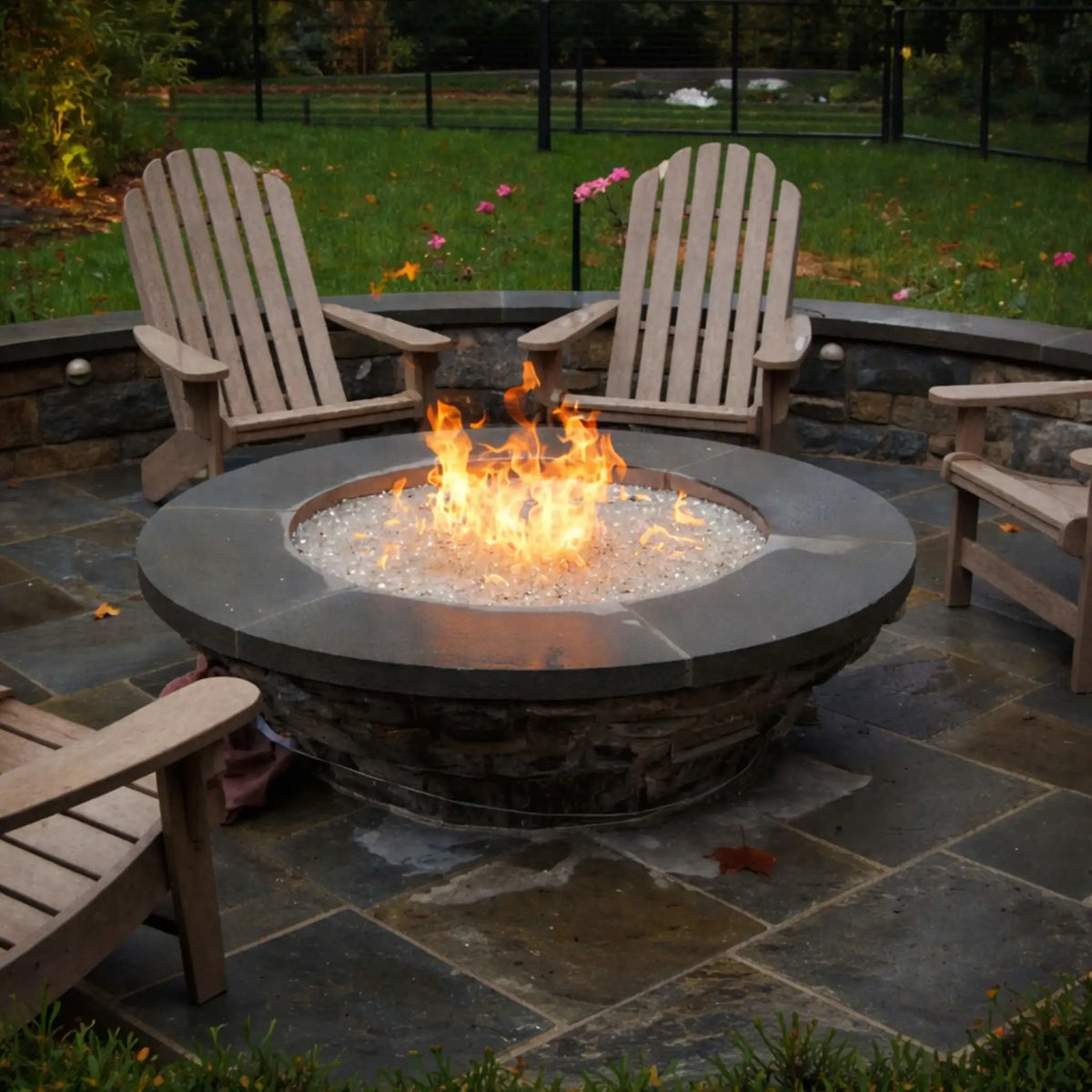 Round outdoor gas fire pit with a dark stone base and smooth concrete top, filled with ultra-clear fire glass and bright flames, surrounded by wooden Adirondack chairs on a wet stone patio, creating a cozy backyard gathering space with lush greenery and garden views.