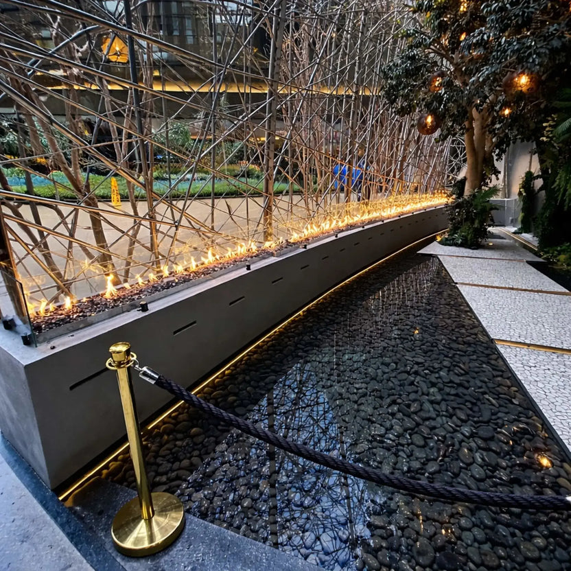 Backyard patio dining area featuring a linear fire pit powered by HPC Fire Hearth Products Torpedo Linear burner system, with lava rock media, steady flames, and a relaxed outdoor setting with table, chairs, and umbrella near a pool.