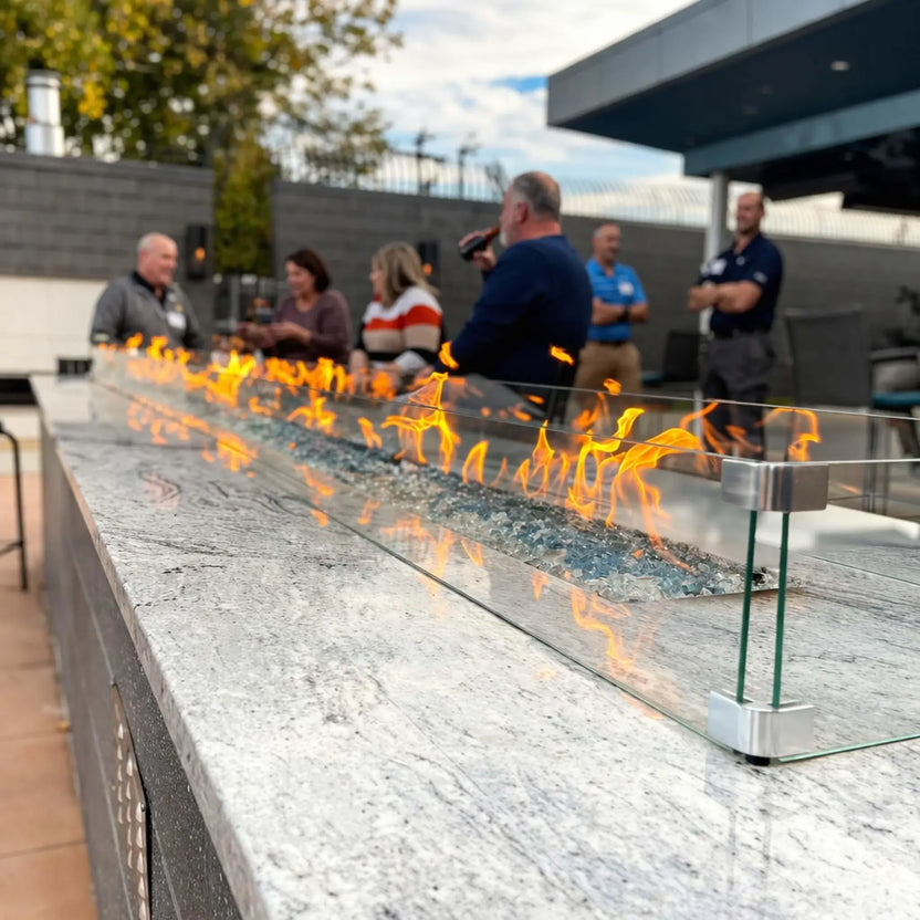 Outdoor social setting featuring a long linear fire table powered by HPC Fire Hearth Products Torpedo Linear Interlink Pan and T Burner, with bright flames dancing over fire glass and a glass wind guard, while guests gather around enjoying the warmth in a modern patio environment.
