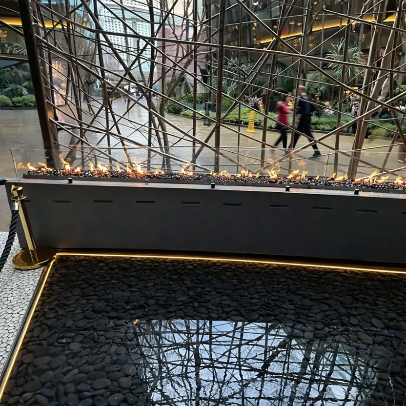 Long straight linear fire feature powered by HPC Fire Hearth Products Torpedo Linear burner system, with continuous flame over lava rock and glass wind guard, set above a reflective water basin and framed by decorative metal branch structures in a high-end indoor setting.