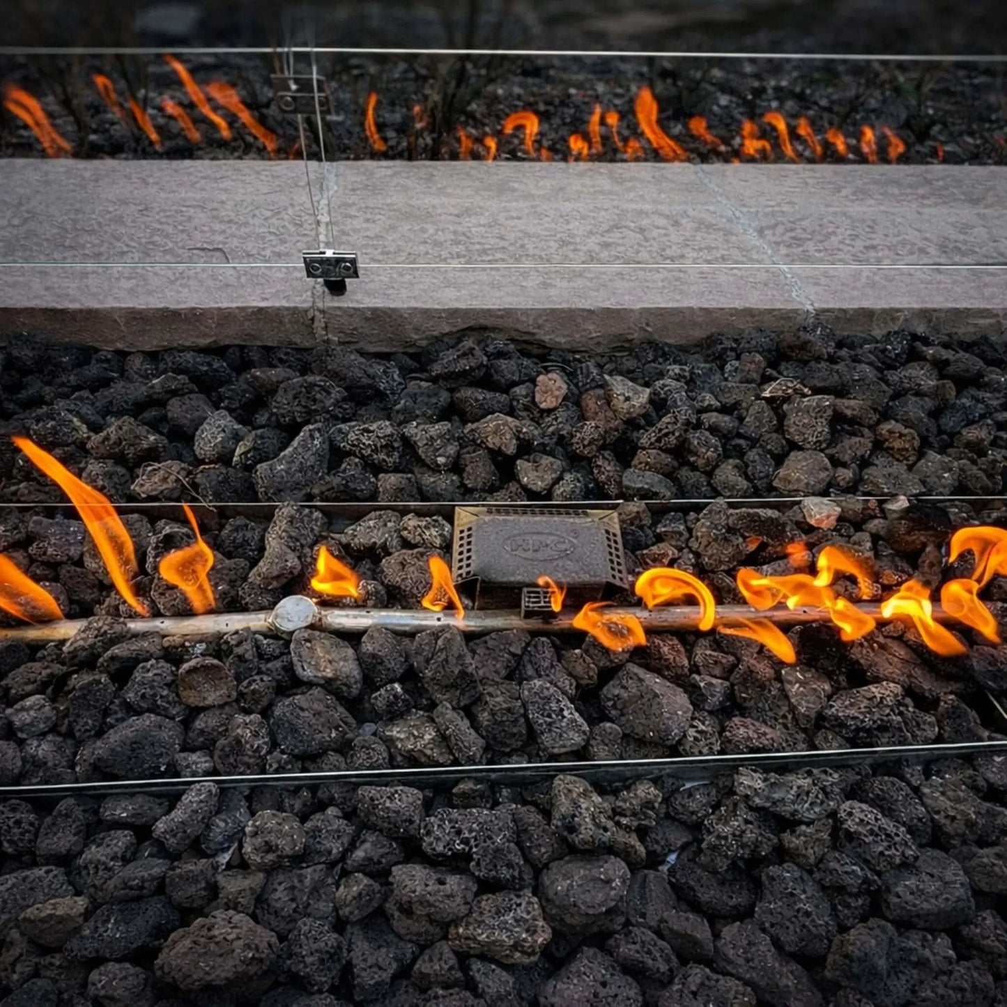 Close-up view of the HPC Fire Torpedo Linear Interlink FPPK burner system installed beneath lava rock media, showing a steady linear flame pattern along the burner tube. The image highlights the push button flame sensing ignition system and even gas distribution within a glass wind guard enclosure.