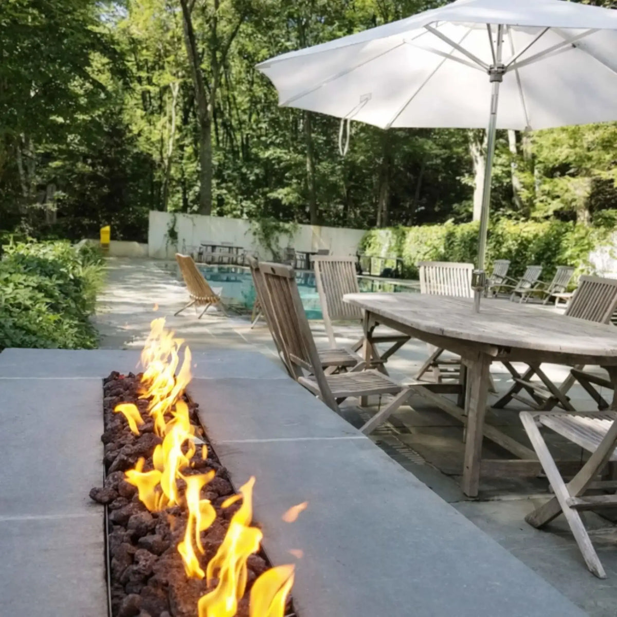 Long linear fire feature built into a stone retaining wall, showcasing the HPC Fire Torpedo Linear Interlink FPPK burner system. Flames stretch across the full length of the installation, enhancing the texture of natural stone and surrounding landscape design.