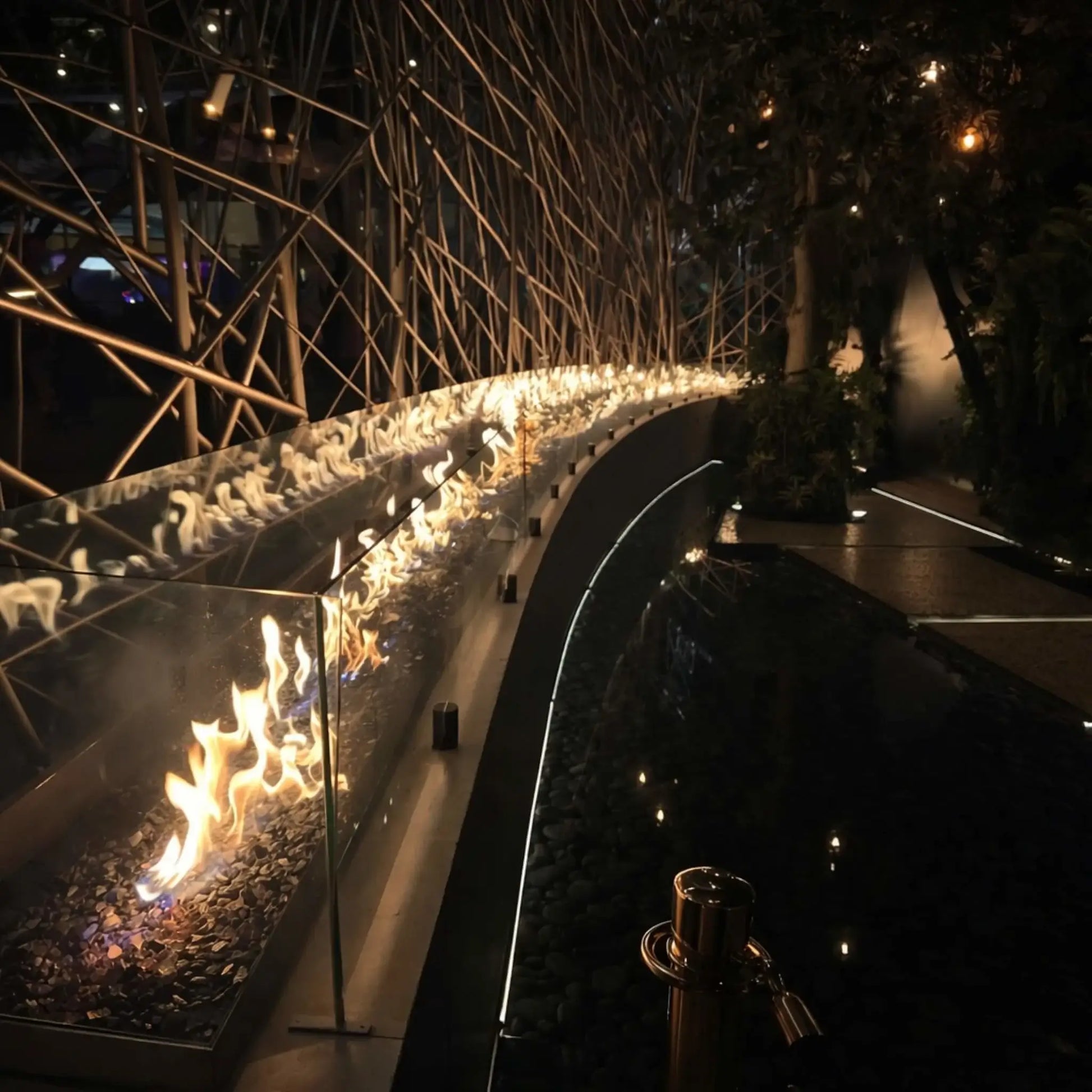 Evening shot of a curved commercial fire installation using HPC Fire Hearth Products Controls torpedo linear interlink pan and T burner system. Flames reflect in water below while a glass wind guard protects the fire, creating a dramatic ambient lighting effect.