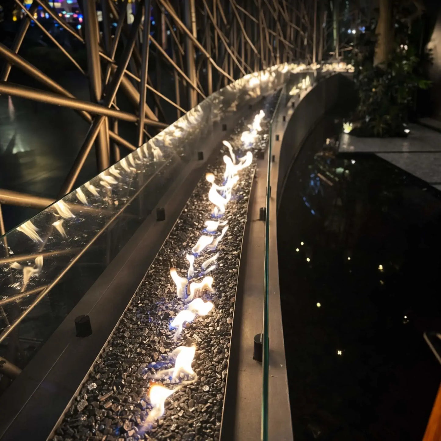 Close-up perspective of HPC Fire Hearth Products Controls torpedo linear interlink burner flames running along a curved fire feature with fire glass media. The glass wind guard and reflective surfaces enhance the visual depth of the high capacity match lit ignition system.