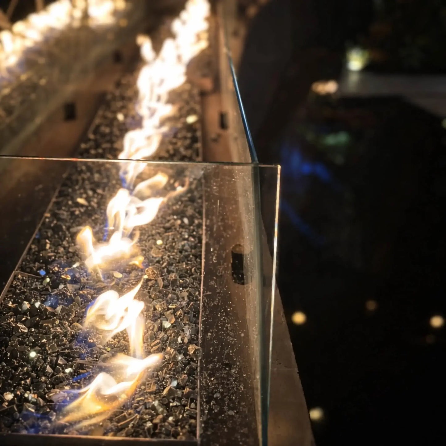 Close-up view of HPC Fire Hearth Products Controls torpedo linear interlink pan and T burner with high capacity match lit ignition, showing bright flames dancing over dark fire glass. A clear glass wind guard lines the edge, while reflections shimmer in nearby water, emphasizing the sleek, modern fire feature design.