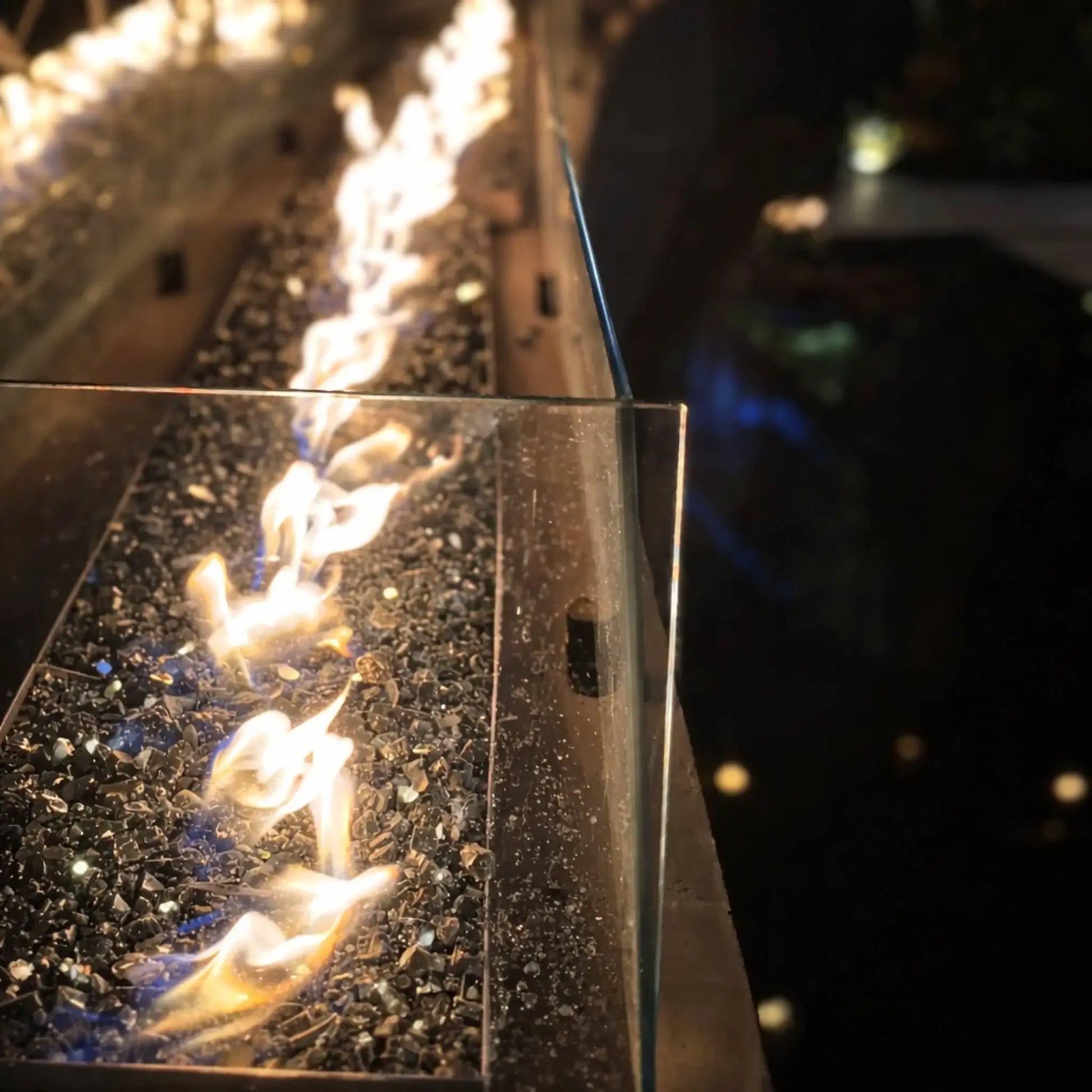 Close-up view of HPC Fire Hearth Products Controls torpedo linear interlink pan and T burner with high capacity match lit ignition, showing bright flames dancing over dark fire glass. A clear glass wind guard lines the edge, while reflections shimmer in nearby water, emphasizing the sleek, modern fire feature design.