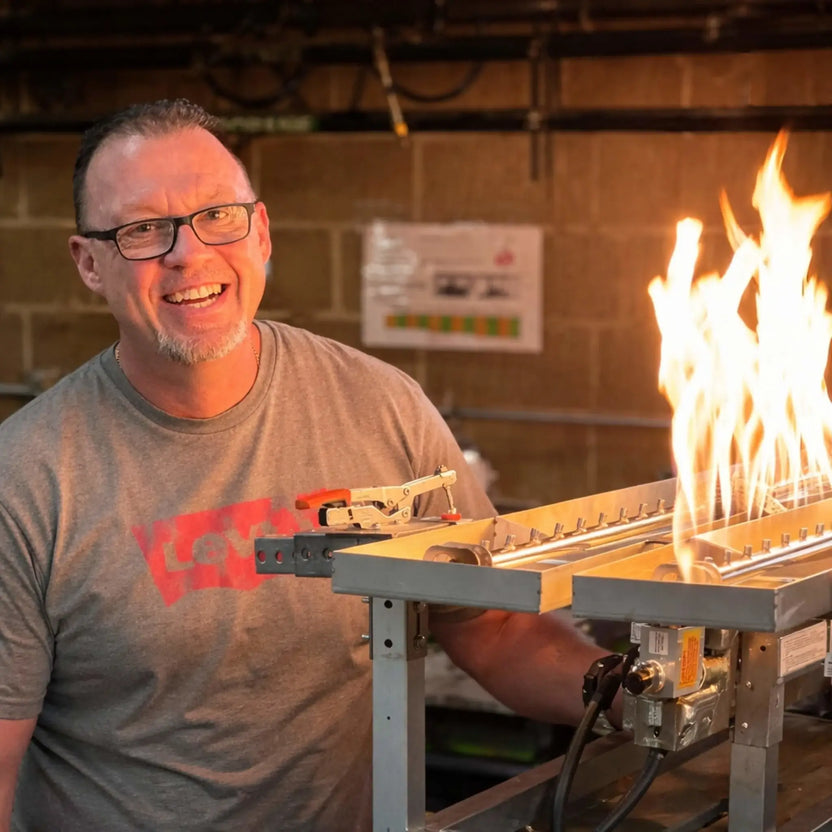 Smiling man demonstrating HPC Fire Hearth Products Controls torpedo linear interlink pan and T burner system indoors, showing active flames rising from the burner. The high capacity match lit ignition setup is mounted on a test stand, emphasizing performance and flame height control.