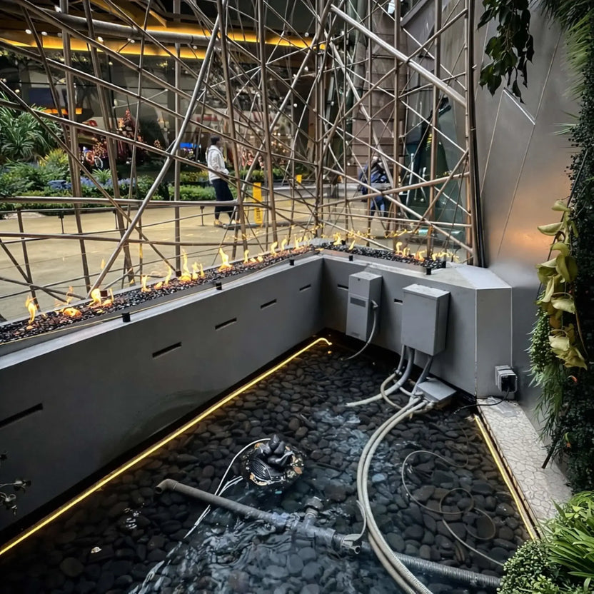 Commercial installation of HPC Fire Hearth Products Controls torpedo linear interlink burner system positioned above a shallow water bed filled with black stones. Visible plumbing and control boxes beneath the feature reveal the functional setup behind the continuous linear flame display.