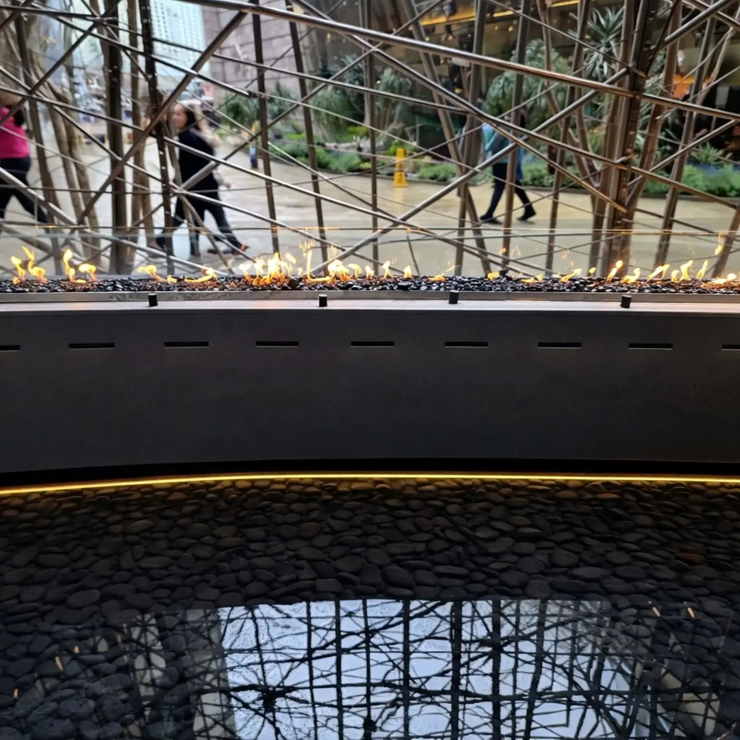 Front-facing view of HPC Fire Hearth Products Controls torpedo linear interlink burner system integrated into a curved fire feature. Flames stretch evenly across the length, while warm LED lighting beneath highlights smooth river stones and reflective water below.