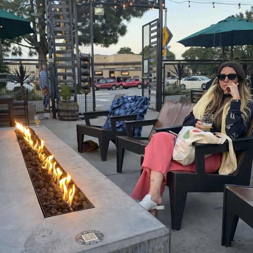 Outdoor patio seating area featuring HPC Fire Hearth Products Controls torpedo linear interlink drop in pan and T burner system built into a concrete fire table. A woman relaxes nearby as a long row of flames burns steadily through lava rock, showcasing real-world lifestyle use.