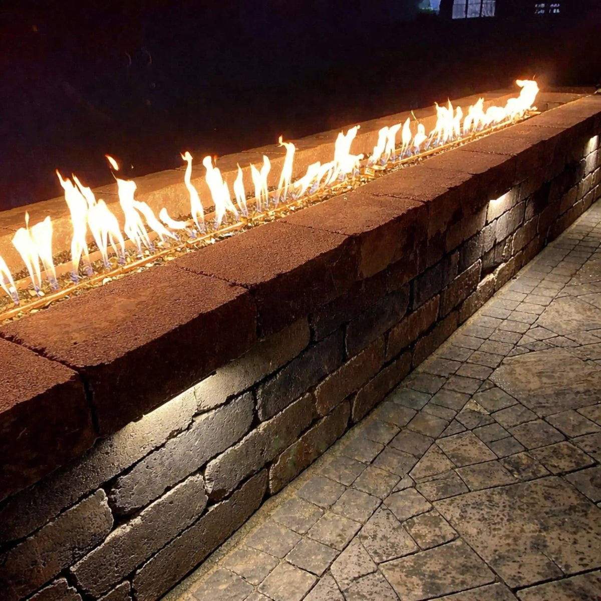 Long outdoor linear gas fire feature built into a textured stone wall, showcasing a continuous ribbon flame over reflective fire glass. Integrated under-cap LED lighting highlights the stacked stone façade, while the surrounding paver patio adds depth and contrast, creating a warm, modern ambiance for nighttime outdoor entertaining.