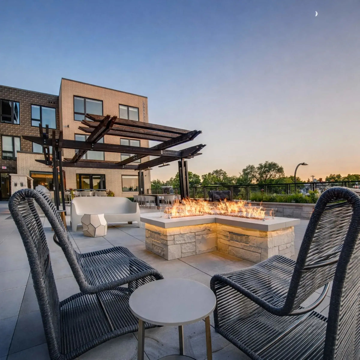 Contemporary rooftop lounge with a square linear fire pit table featuring a bright flame pattern over fire media. Surrounded by modern woven lounge chairs and a sleek pergola structure, the setting overlooks a landscaped urban backdrop, blending clean architectural lines with inviting outdoor comfort.