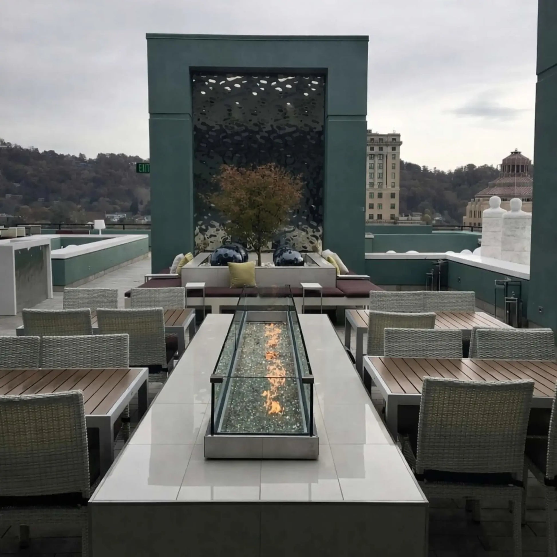 Rooftop lounge area with a long rectangular fire table featuring a linear burner and glass wind guard. The fire glows over reflective fire glass, surrounded by cushioned seating and woven outdoor furniture. A decorative water wall and cityscape backdrop enhance the upscale, modern outdoor gathering space.