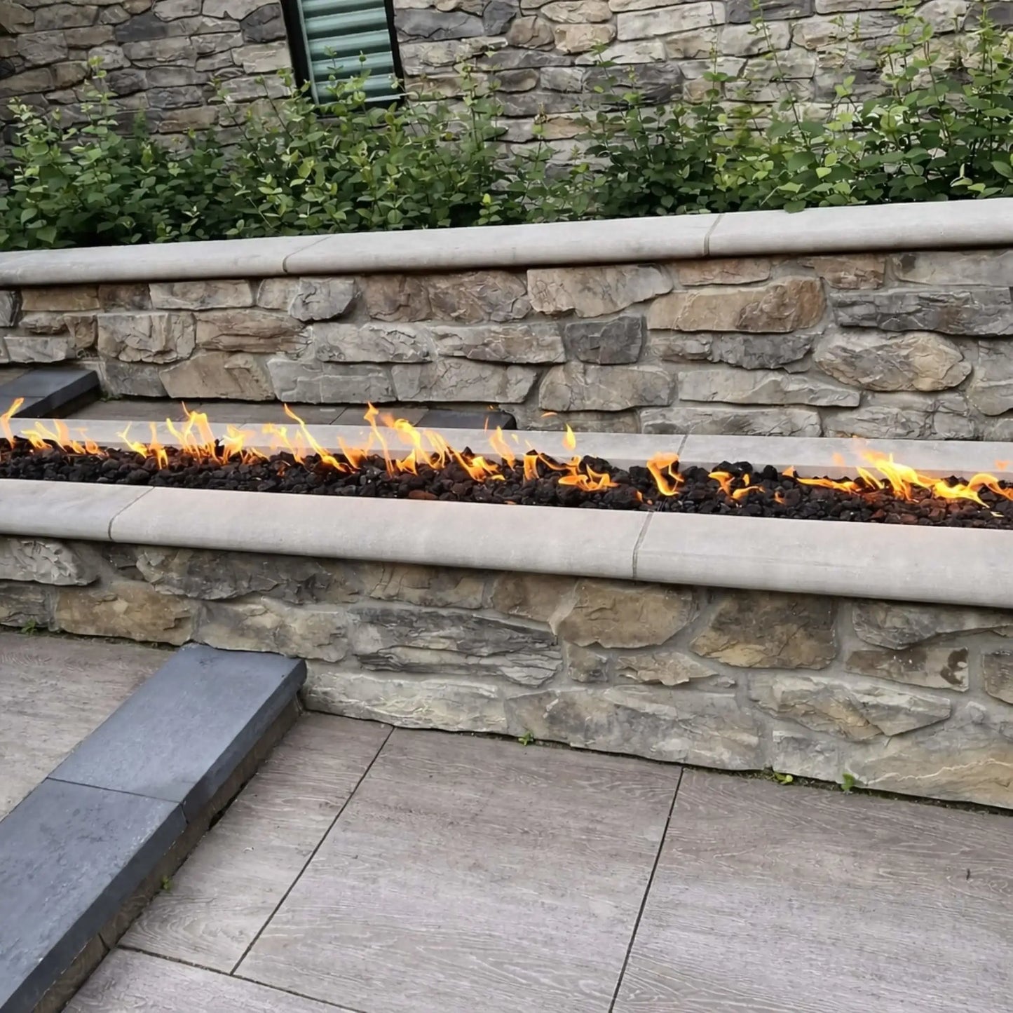 Outdoor stacked stone fire wall featuring a built-in linear gas burner with bright flames running across black lava rock. The clean stone cap and surrounding landscaping create a polished, modern hardscape design, with steps and patio surfaces framing the fire feature in a residential backyard.