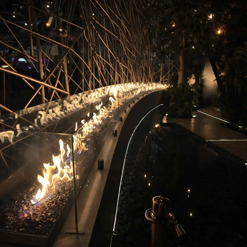 Wide-angle view of a curved indoor fire and water feature with a long linear burner producing warm flames along a glass-lined channel. The installation follows a sweeping architectural line next to a reflective pool filled with dark stones, surrounded by sculptural metal framework and soft ambient lighting.