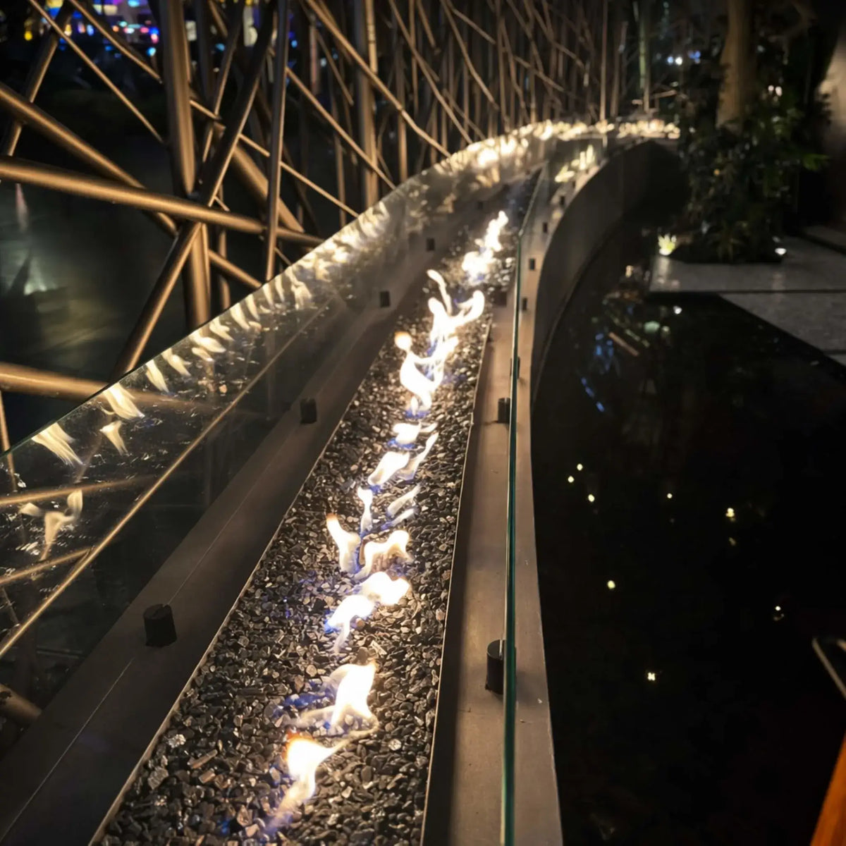 Curved indoor linear fire feature with a continuous flame line over black fire glass, protected by a glass wind barrier. The sleek metal burner runs alongside a reflective water basin, framed by an intricate metal lattice structure, creating a dramatic architectural fire and water installation in a dimly lit space.