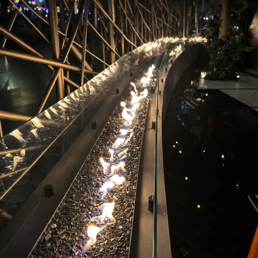 Curved indoor linear fire feature with a continuous flame line over black fire glass, protected by a glass wind barrier. The sleek metal burner runs alongside a reflective water basin, framed by an intricate metal lattice structure, creating a dramatic architectural fire and water installation in a dimly lit space.