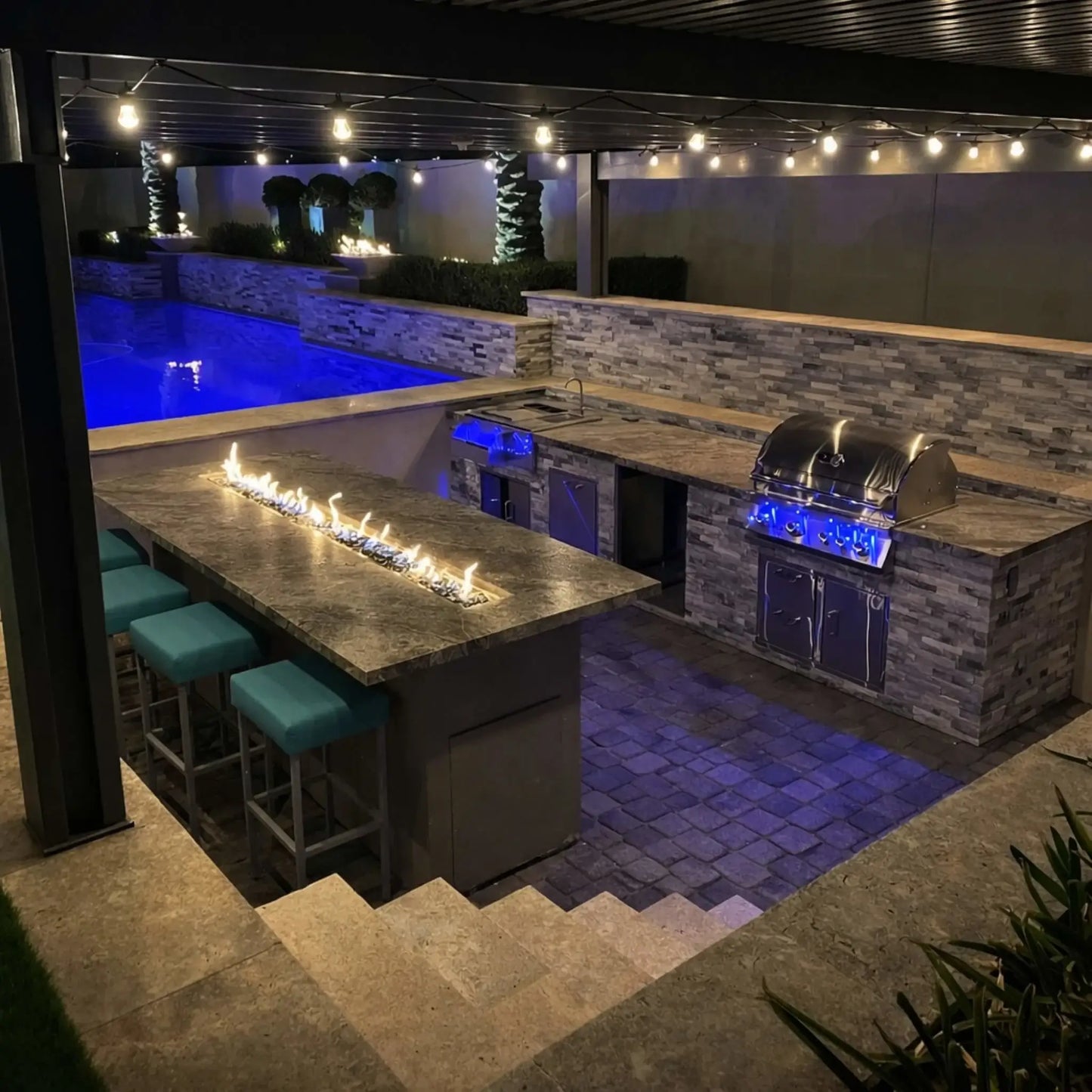 Luxury outdoor kitchen and bar featuring a long linear gas fire table with a continuous flame strip set into a stone countertop. Teal bar stools line the seating area, while a built-in stainless steel grill and sink sit beneath warm string lighting. A glowing blue pool and raised stone fire features in the background create a high-end backyard entertainment space.