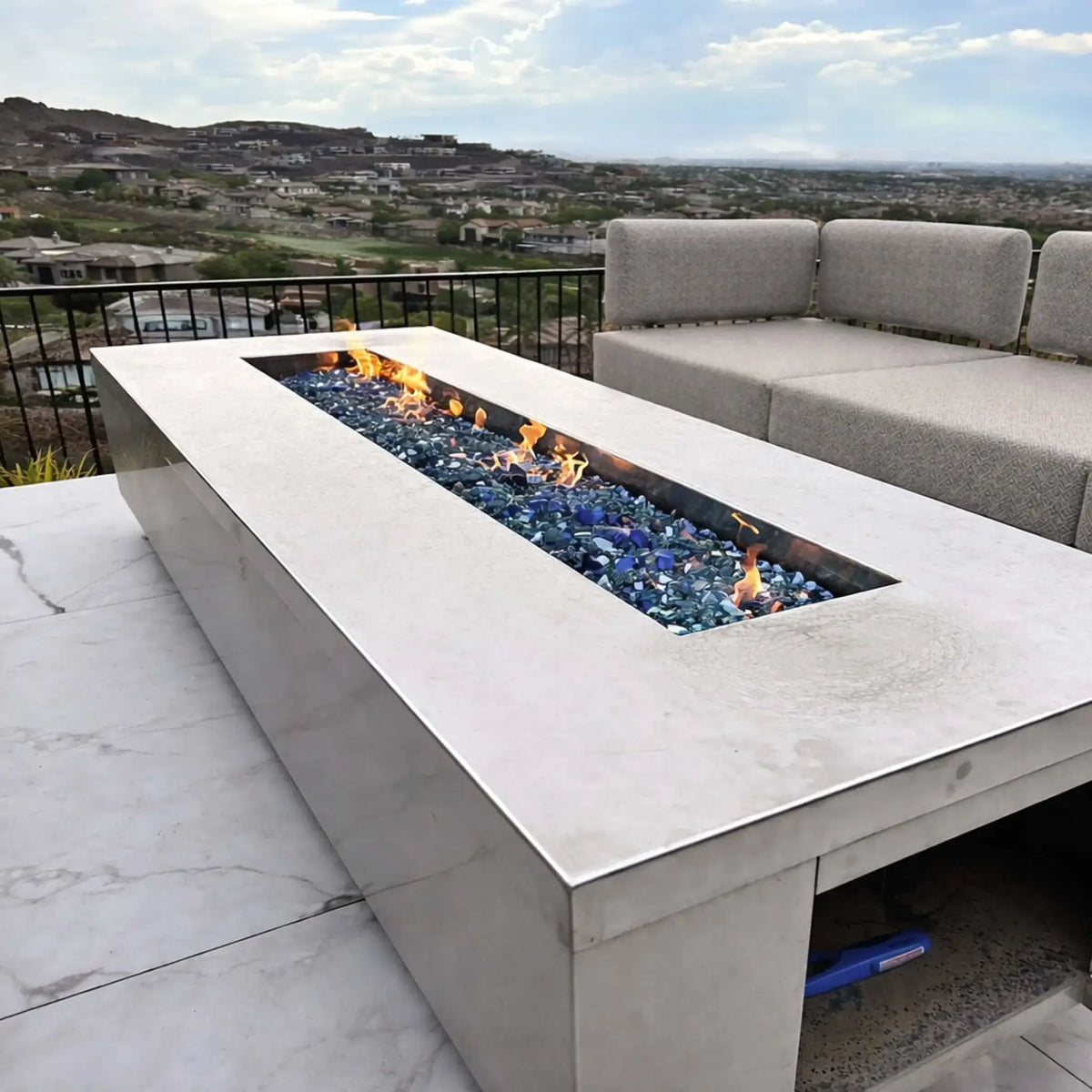 Contemporary rectangular fire pit table with a recessed linear burner filled with blue fire glass, producing a clean flame pattern. The unit sits on a rooftop patio overlooking a suburban hillside neighborhood, paired with modern outdoor seating for a relaxed, upscale outdoor living space.