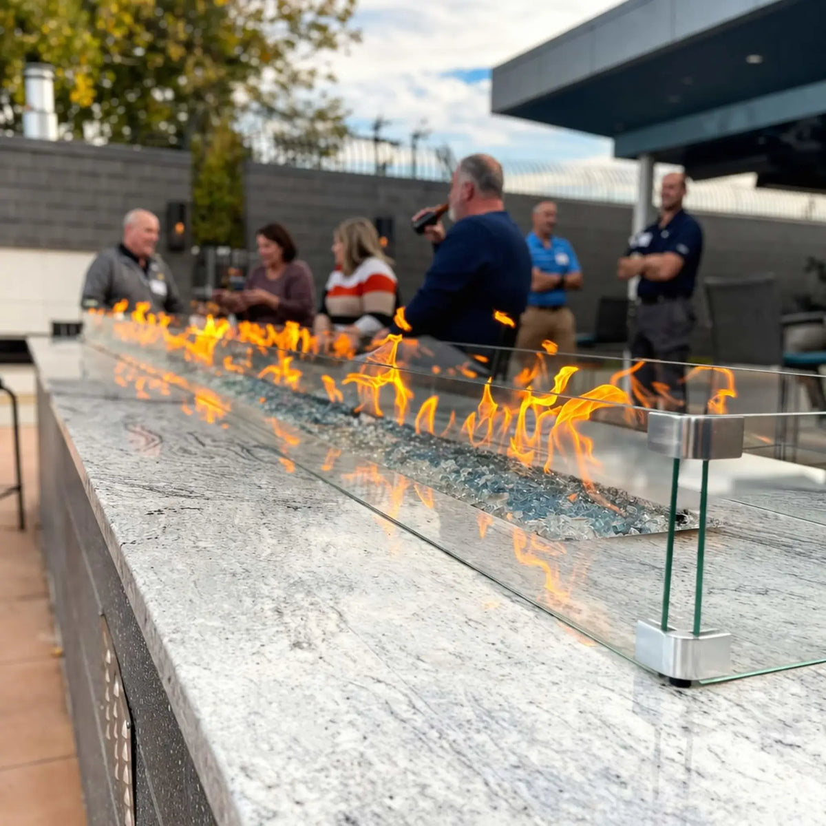 Outdoor linear fire table with clear glass wind guard and blue fire glass media, creating vibrant flames along a polished stone surface. A group of people gathers around the fire feature in a modern patio setting, highlighting warmth, ambiance, and social interaction.