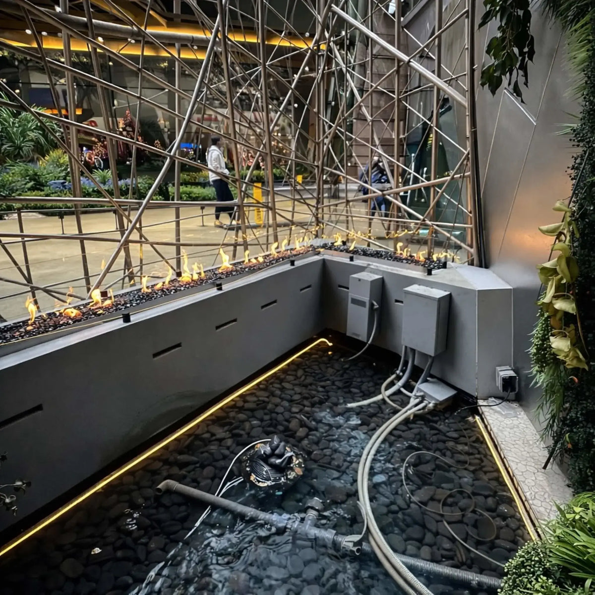 Corner installation of a linear fire feature integrated with a water basin, showing exposed plumbing, control boxes, and submerged components beneath the burner. Flames dance above dark fire glass, while the surrounding modern structure includes metal lattice elements and indoor landscaping.