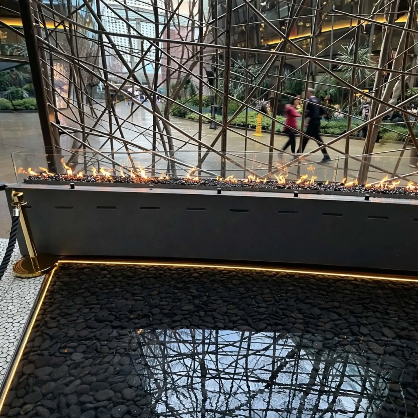 Wide view of a contemporary indoor fire and water feature combining a linear gas burner and reflective water basin filled with black stones. The fire runs along a sleek metal enclosure beneath a sculptural metal framework, with people walking through a modern, open-air atrium behind it.