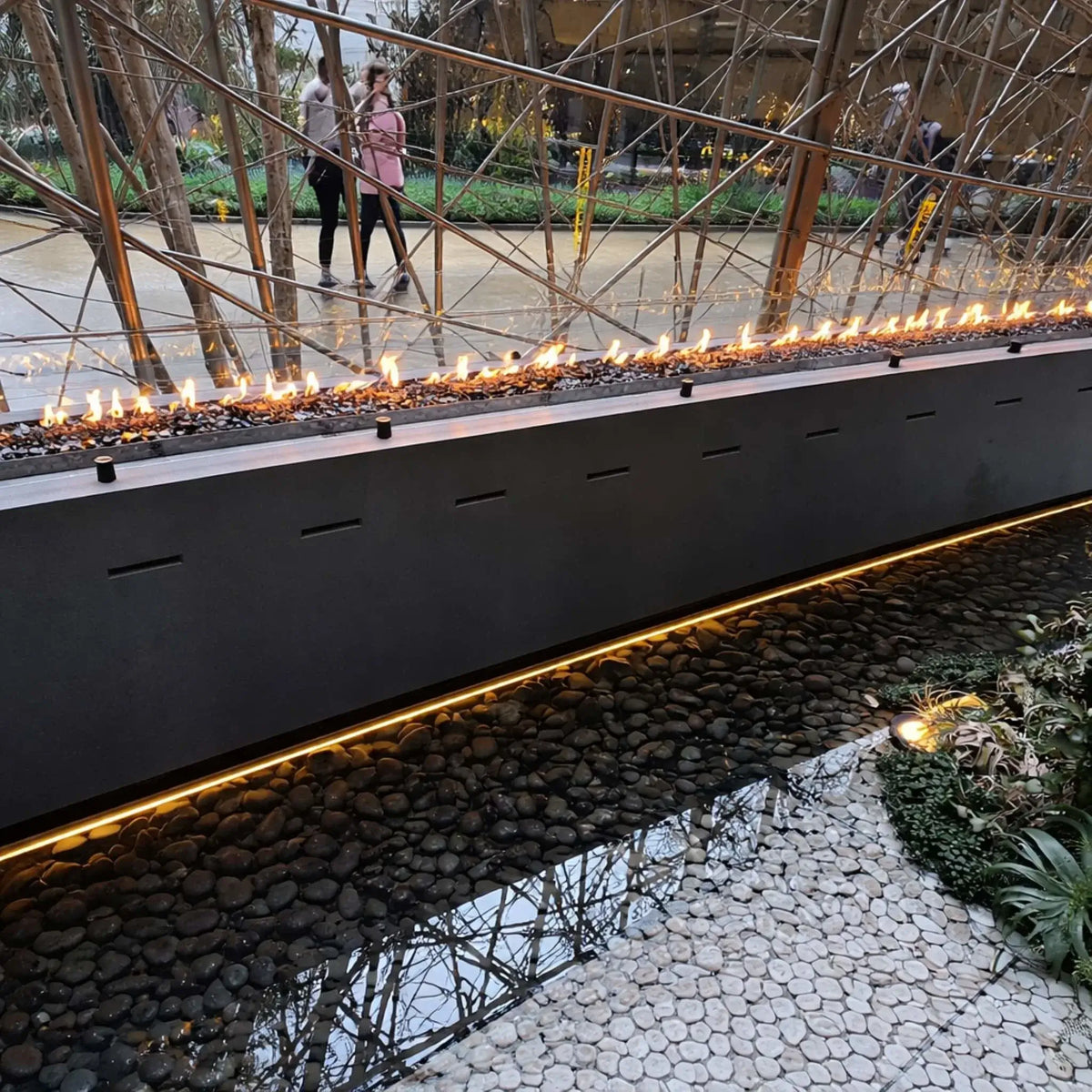 Modern indoor linear fire feature with a sleek black metal housing and continuous ribbon flame over dark fire glass. The burner runs alongside a reflective water basin filled with smooth black river rocks, set within a contemporary architectural space featuring metal branch-like structures and landscaped greenery in the background.
