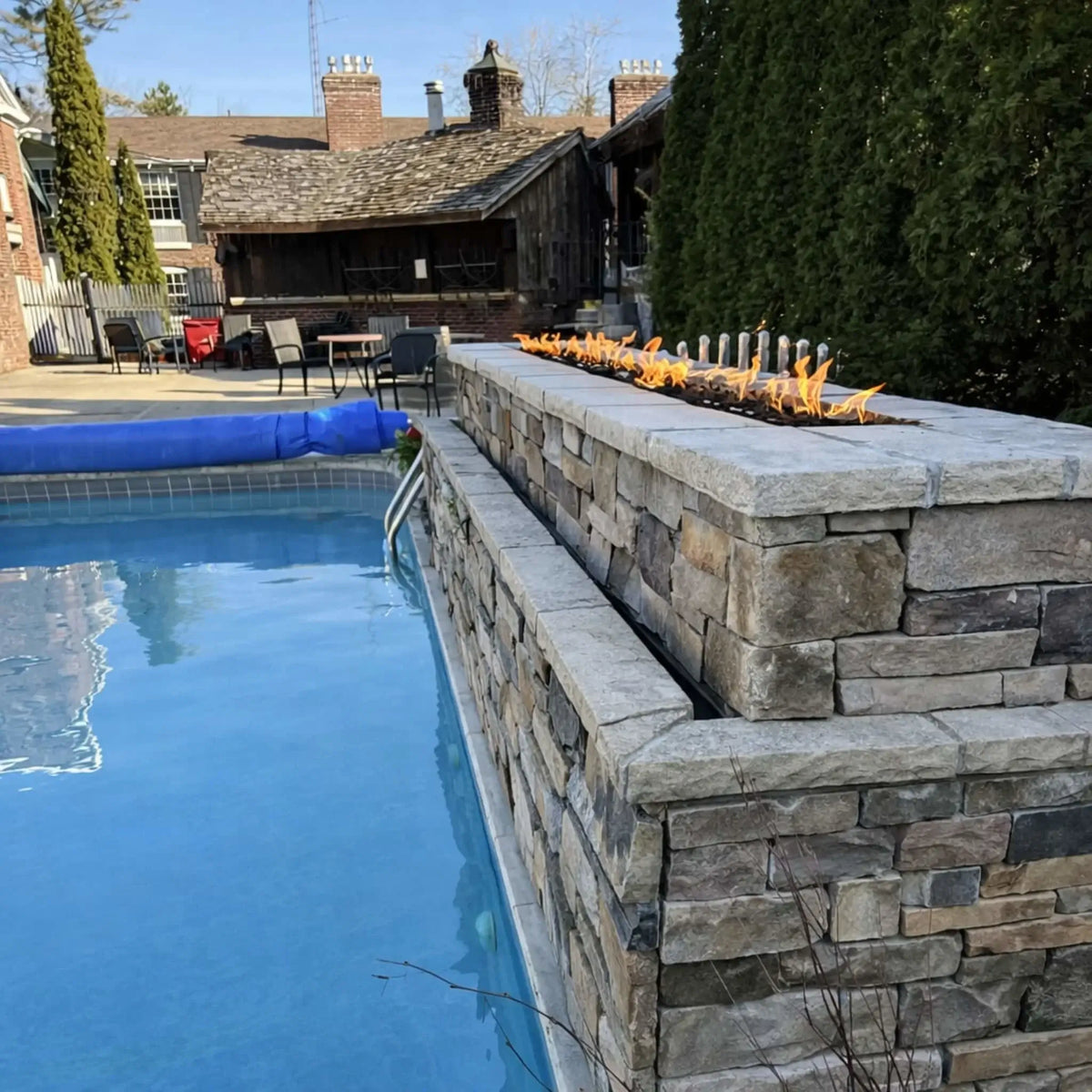 Outdoor poolside stone fire wall with a long linear gas burner producing a clean flame line above dark fire media. The stacked stone structure borders a swimming pool, with patio seating and residential buildings in the background, creating a warm, inviting backyard entertainment setting.