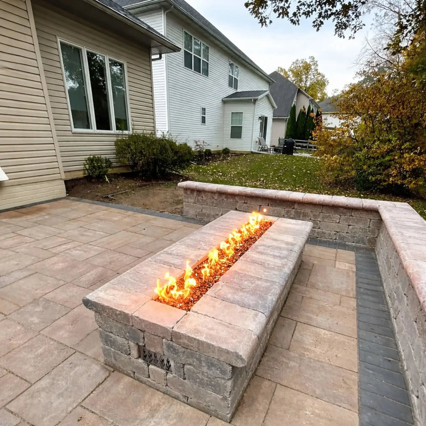 Residential backyard installation showcasing a rectangular stone fire pit powered by the HPC Fire Torpedo Linear Interlink MLFPK burner system. The linear flame runs through fire media in a paver patio setting, surrounded by retaining walls, lawn space, and a suburban home exterior.