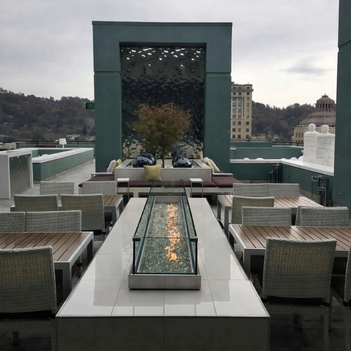Modern rooftop lounge featuring a sleek linear fire table with glass wind guard, powered by the HPC Fire Torpedo Linear Interlink MLFPK burner system. The flame runs through fire glass media, surrounded by contemporary seating and cityscape views, emphasizing upscale outdoor design.