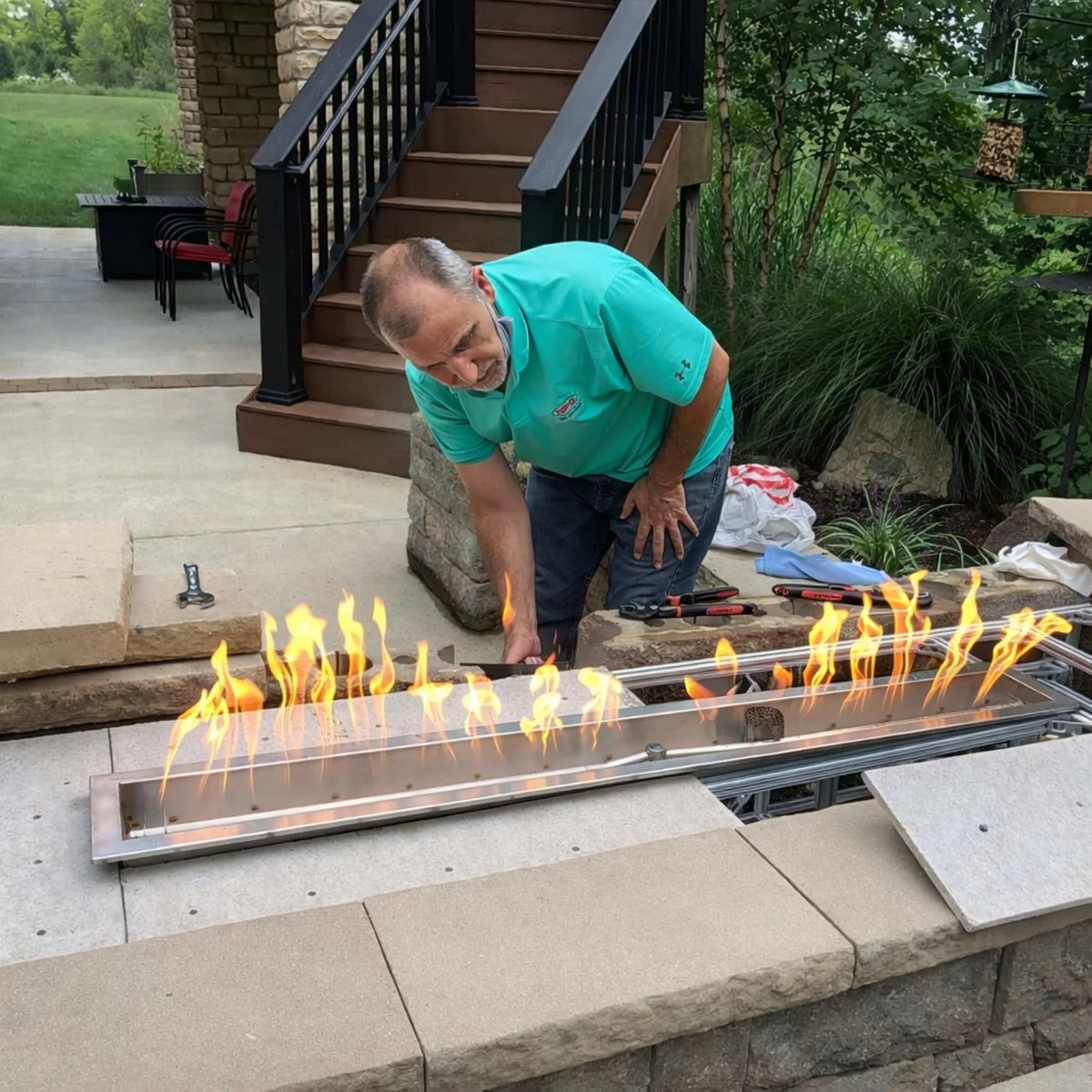 Installer adjusting an HPC Fire Torpedo linear trough pan and T-burner system during setup in a backyard stone fire feature. Flames are actively burning at a mid-to-high Hi setting, showcasing real-world installation and the Hi/Lo flame control capability using electronic ignition with remote, app, or smart tablet operation.