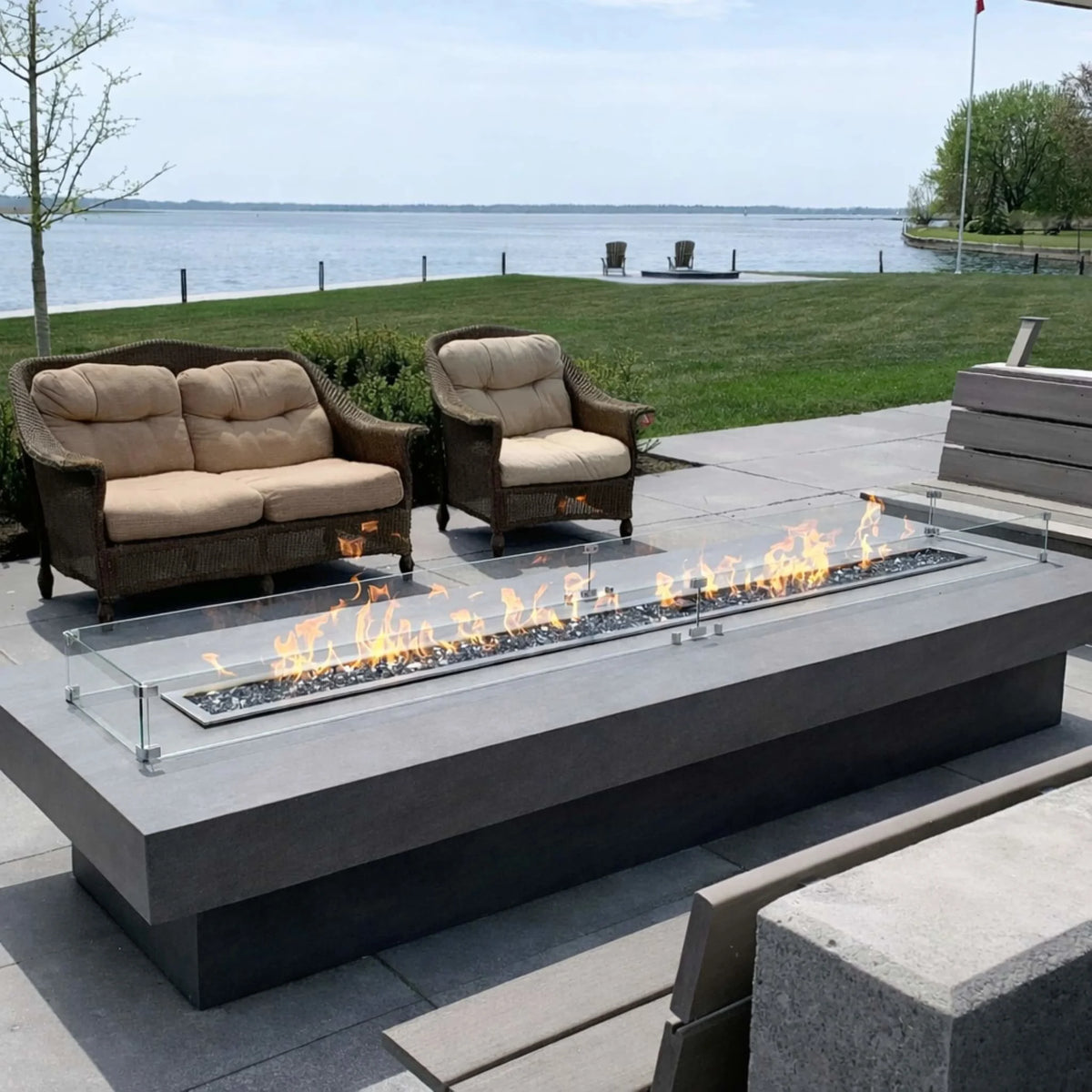Lakeside patio featuring a sleek linear fire table with an HPC Torpedo burner and glass wind guard. Flames stretch evenly across at a controlled medium setting, demonstrating smooth Hi/Lo flame adjustment via electronic ignition, perfect for relaxing outdoor seating near the water.
