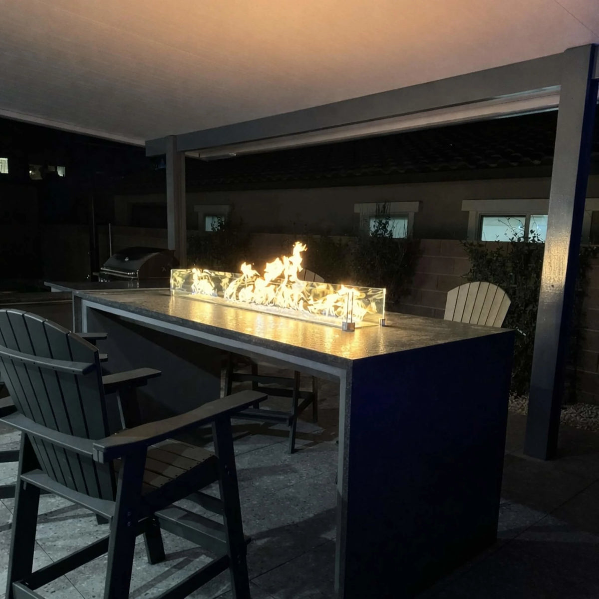 Nighttime outdoor bar setup with a modern fire table using an HPC Torpedo linear burner system. Flames glow brightly at a higher Hi setting behind a glass wind guard, highlighting ambiance and the ease of adjusting flame height with remote or smart device Hi/Lo controls.