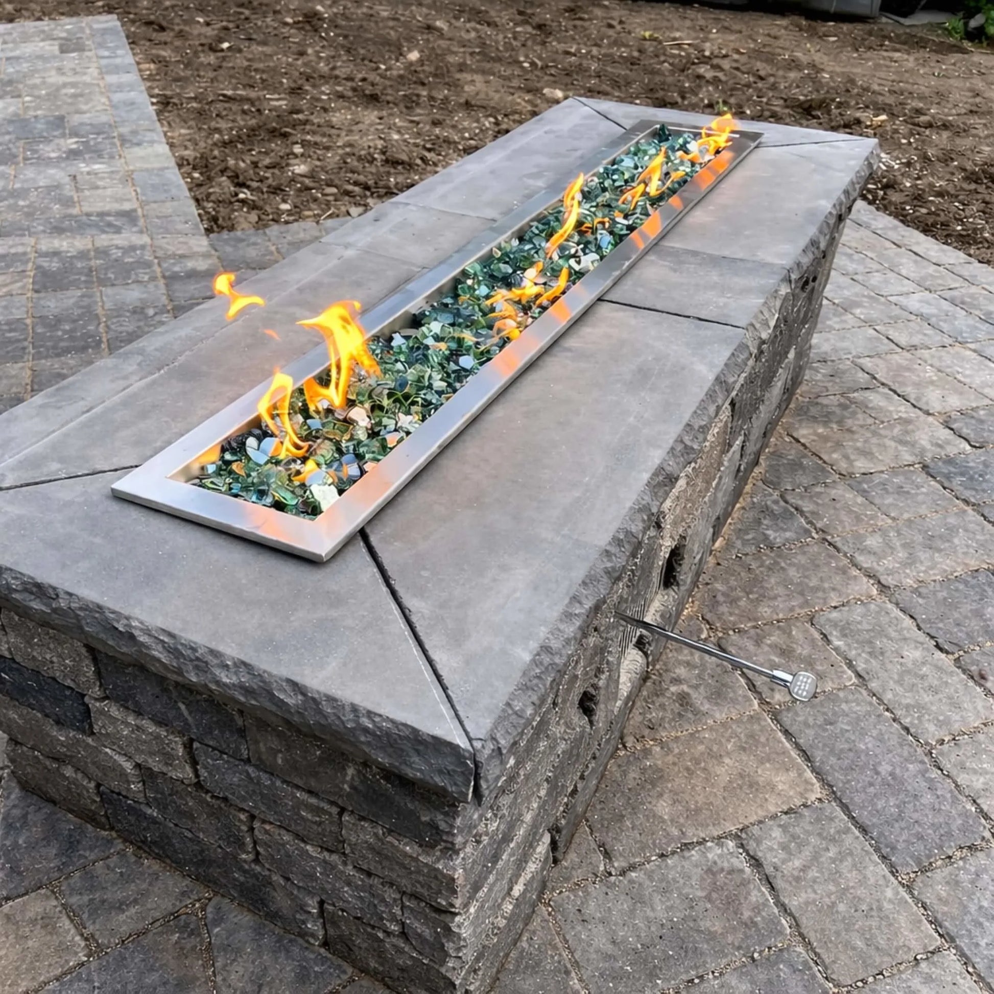 Freshly installed outdoor fire feature with green fire glass and an HPC Torpedo linear trough burner. Flames flicker at a moderate setting, demonstrating adjustable Hi/Lo performance and consistent ignition across the full burner length.
